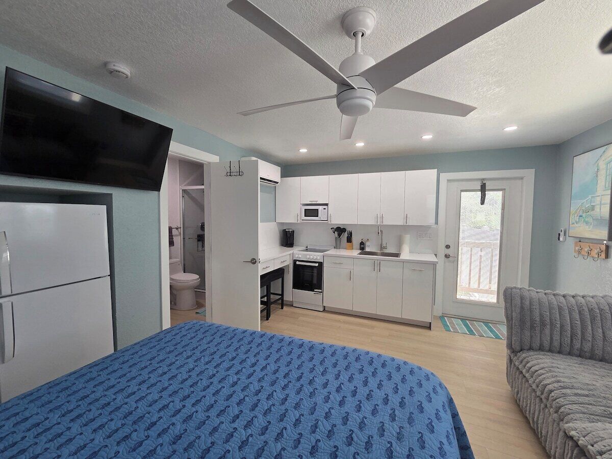 A compact room with a bed, kitchenette, and a ceiling fan.