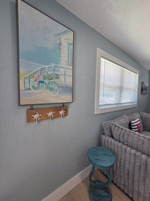 A room with a painting of a bicycle, a window with blinds, a grey sofa, and a small stool. - Ocean Whispers at Tuscany Sun: Beach Studio (Melbourne Beach)