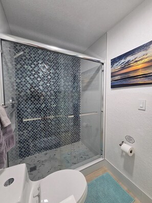 A bathroom with a glass shower enclosure, a white sink, a toilet paper holder, and a painting on the wall. - Ocean Whispers at Tuscany Sun: Beach Studio (Melbourne Beach)