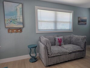 A room with a grey sofa, a small round table, a window with blinds, and wall art.