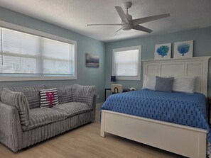 A bedroom with a bed, a gray sofa with a decorative pillow, a ceiling fan, and two windows with blinds.