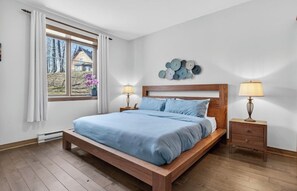 A bedroom with a wooden bed, two bedside tables with lamps, a window with curtains, and a wall decoration. - Cozy Tremblant Escape| Pool & Hot tub (Mont-Tremblant)