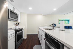 A modern kitchen with stainless steel appliances, a white countertop, and a view of the living area. - Sandcastle Condominium Unit 109 (Port Aransas)
