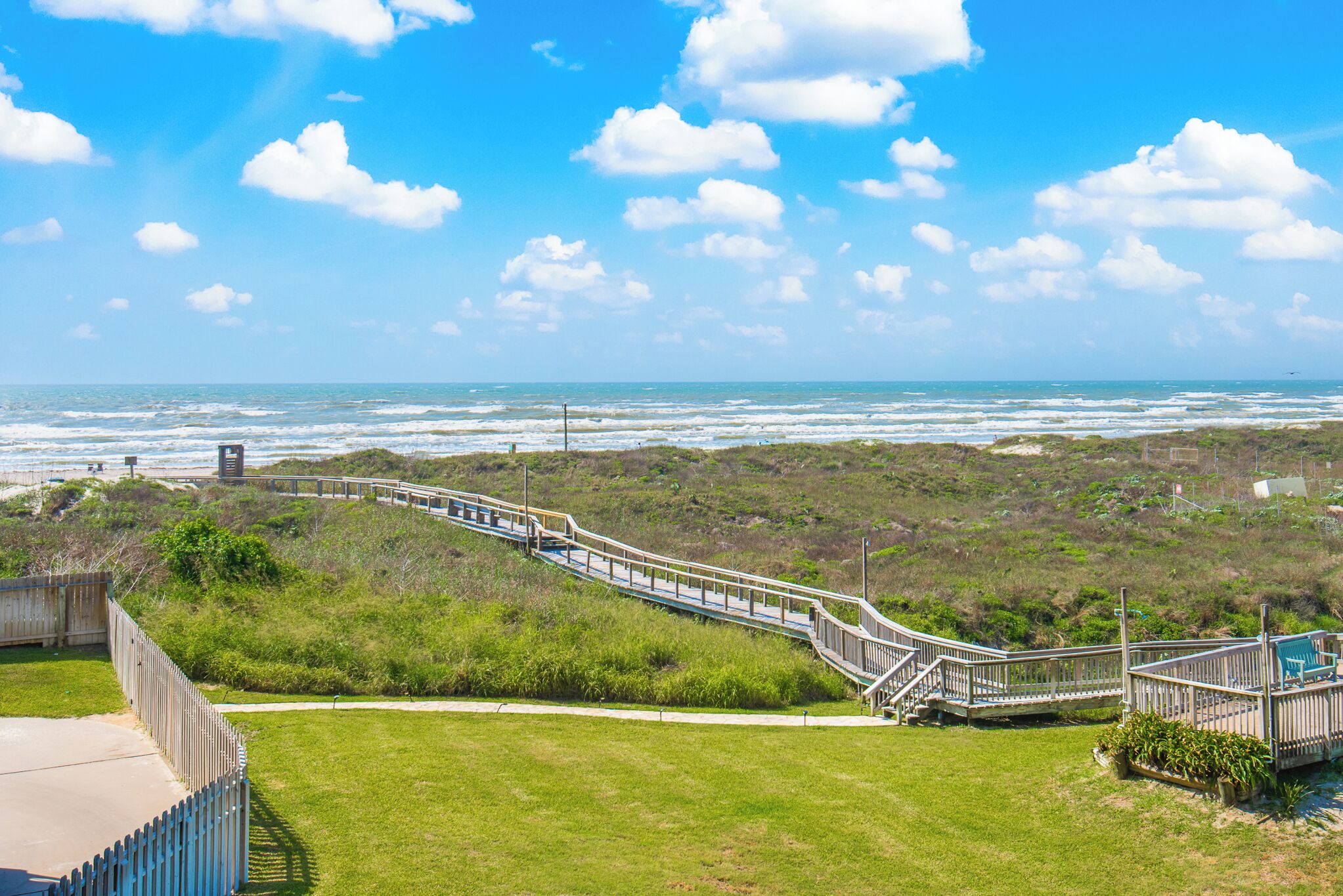 A coastal walkway with a wooden deck, grassy areas, and a view of the ocean.
