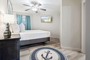 Un dormitorio con cama, escritorio con lámpara, ventilador de techo, cuadro enmarcado y alfombra redonda con diseño de ancla.