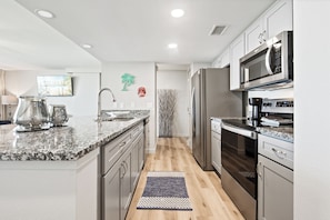 A modern kitchen with granite countertops, stainless steel appliances, and a herringbone-patterned rug. - Sandcastle Condominiums Unit 209 (Port Aransas)