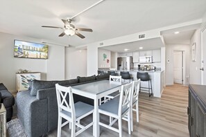 Una sala moderna con un sofá gris, una mesa de comedor blanca con sillas, un ventilador de techo y una cocina con armarios y electrodomésticos.