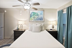 A bedroom with a large bed, two bedside tables with lamps, a ceiling fan, a framed painting, and a window with curtains. - Sandcastle Condominiums Unit 209 (Port Aransas)
