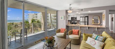 A modern living room with a balcony overlooking a beach, a kitchen with stainless steel appliances, and a cozy seating area with a yellow sofa.