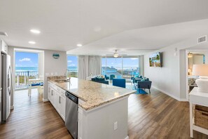 A modern kitchen with a large island, a dining area with blue chairs, and a view of the ocean. - Sandcastle Condominiums 338 (Port Aransas)