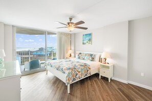 Een slaapkamer met een bed, nachtkastjes, een plafondventilator en een groot raam met uitzicht op het strand en huizen.