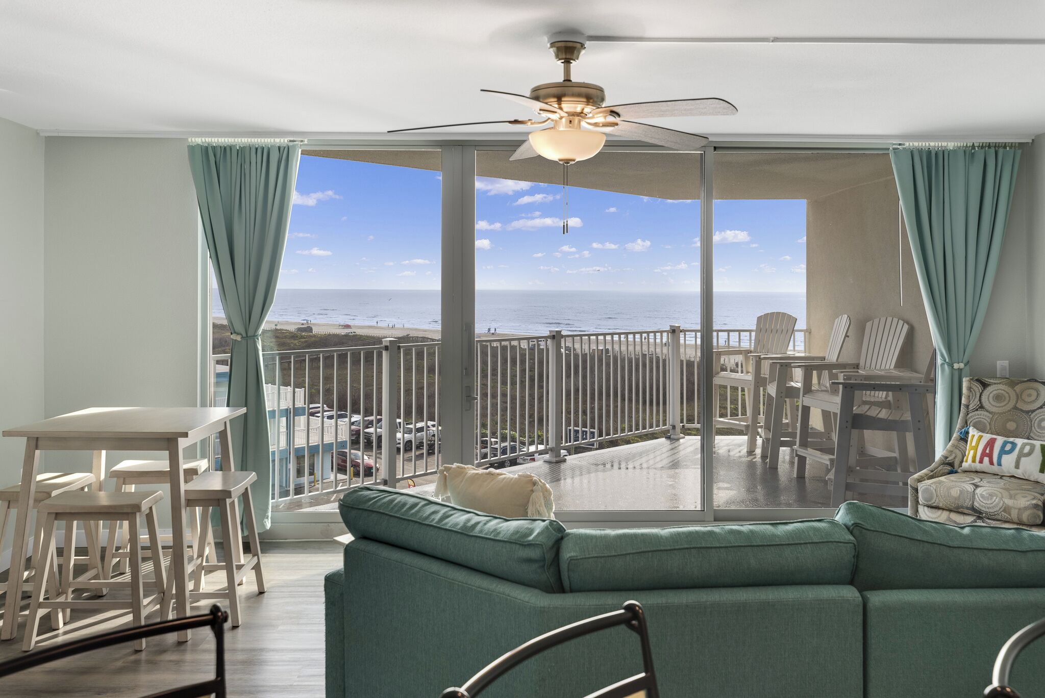 A balcony with a view of the beach, a ceiling fan, and a seating area with a sofa and chairs.