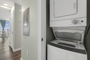 A laundry room with a front-loading washer and dryer, a control panel with various settings, and a view of a balcony with a table and chairs. - Sandcastle Condominiums 408 (Port Aransas)