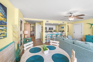 Uma sala de estar ampla com um sofá grande, um espaço de jantar com mesa e cadeiras, e uma cozinha ao fundo.