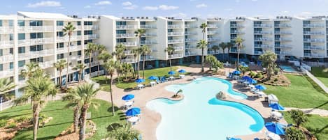 Ett hotell med en pool omgiven av solstolar och parasoller, palmer och en flervåningsbyggnad med balkonger.