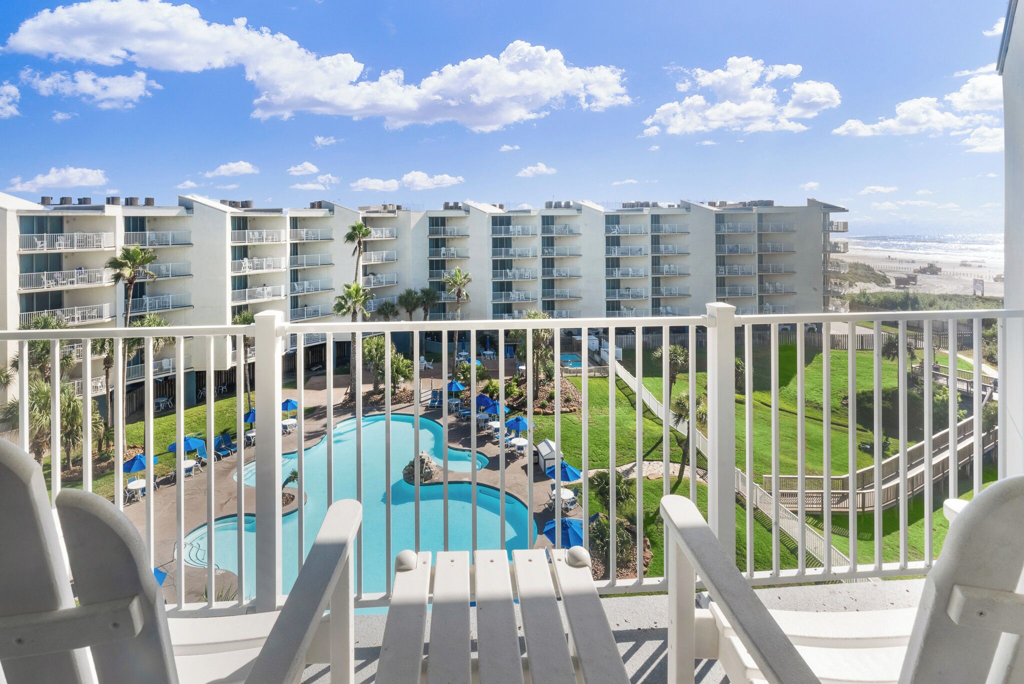 A hotel balcony with a view of a swimming pool, lounge chairs, and a beach.