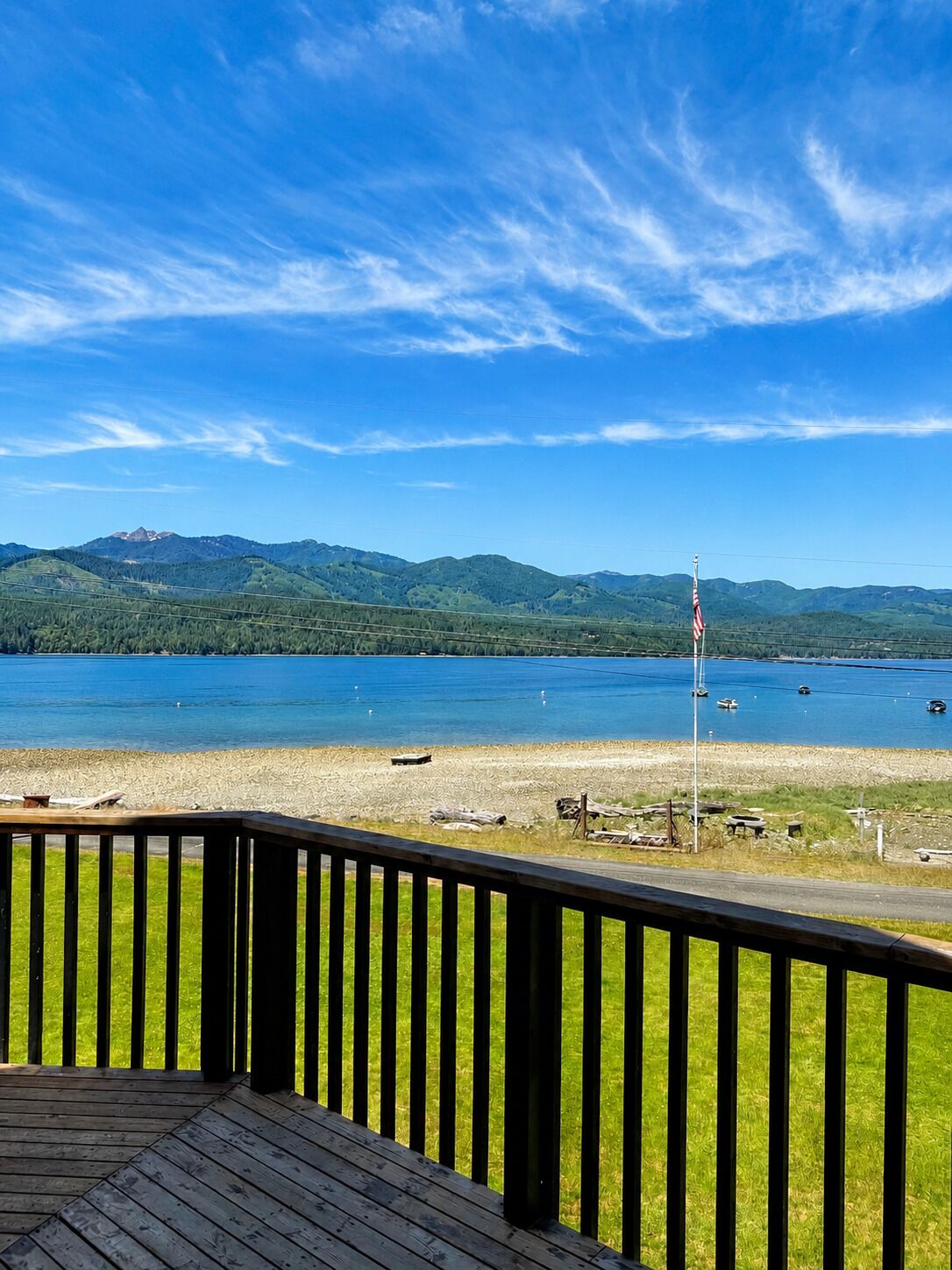 Terraza de madera con vista a un lago con barcos, un área verde y montañas al fondo.