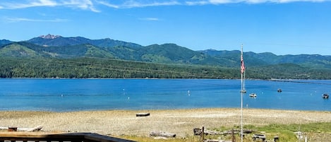 Une terrasse en bois donnant sur un lac où l’on peut apercevoir des bateaux, une zone herbeuse et, au loin, des montagnes.