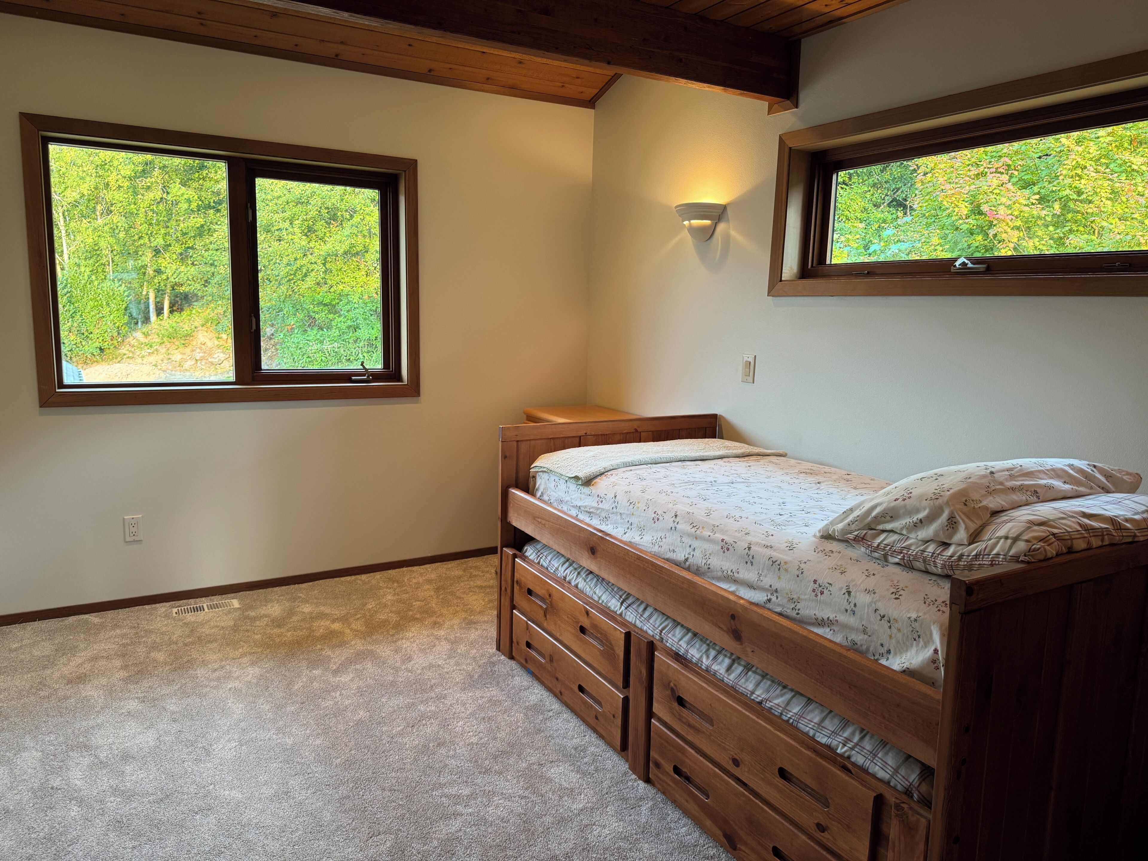 Un dormitorio con literas, dos ventanas con vistas a zonas verdes, una lámpara de pared y suelo alfombrado.