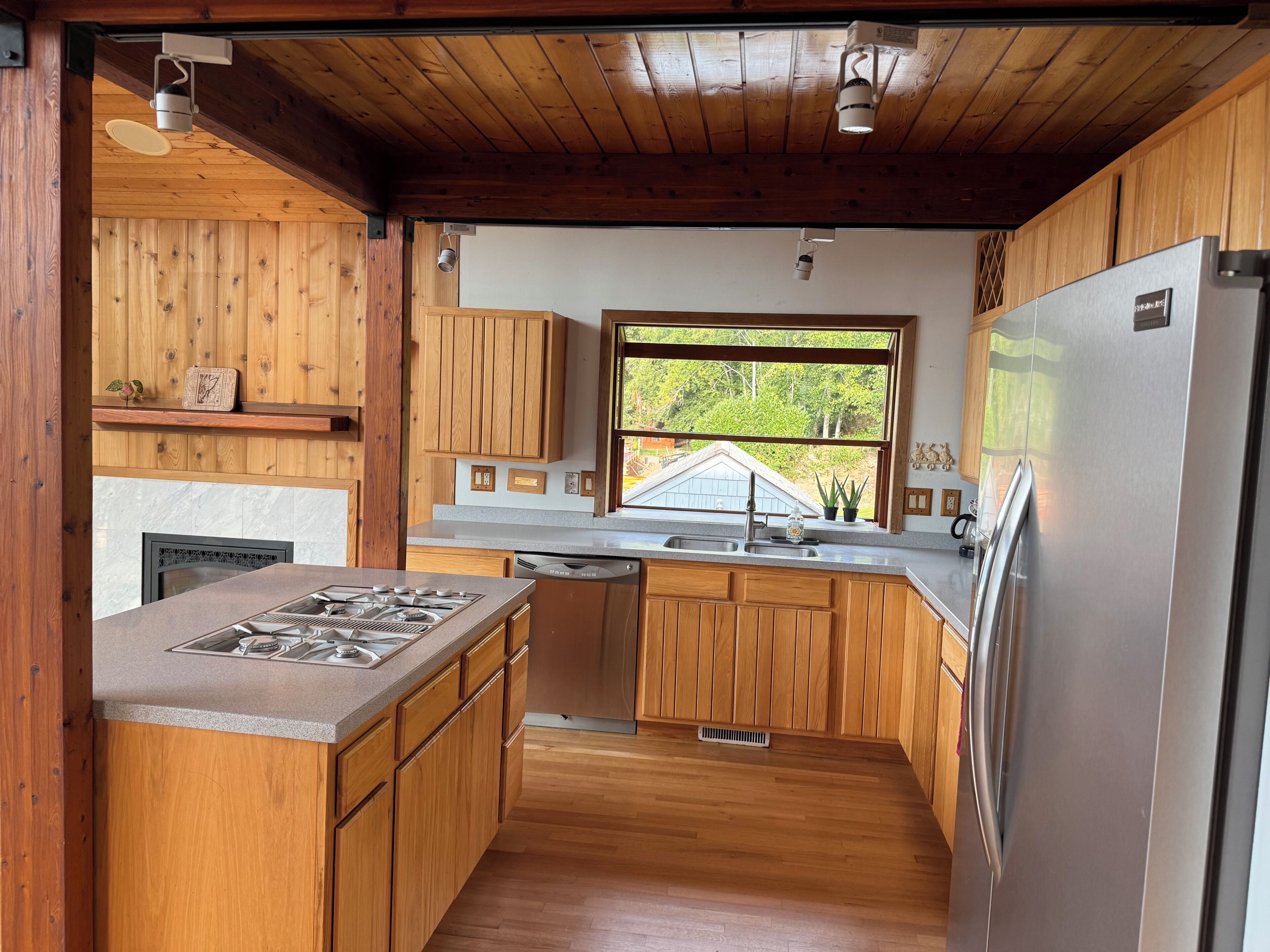 A kitchen with wooden cabinets, a stainless steel refrigerator, and a window with a view of greenery.