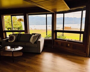 A room with a sofa, a small table, and large windows offering a view of a lake and mountains. - Beachfront Getaway on Hood Canal (Seabeck)