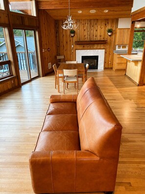 A spacious living area with a leather sofa, wooden flooring, a dining table with chairs, a fireplace, and large windows. - Beachfront Getaway on Hood Canal (Seabeck)