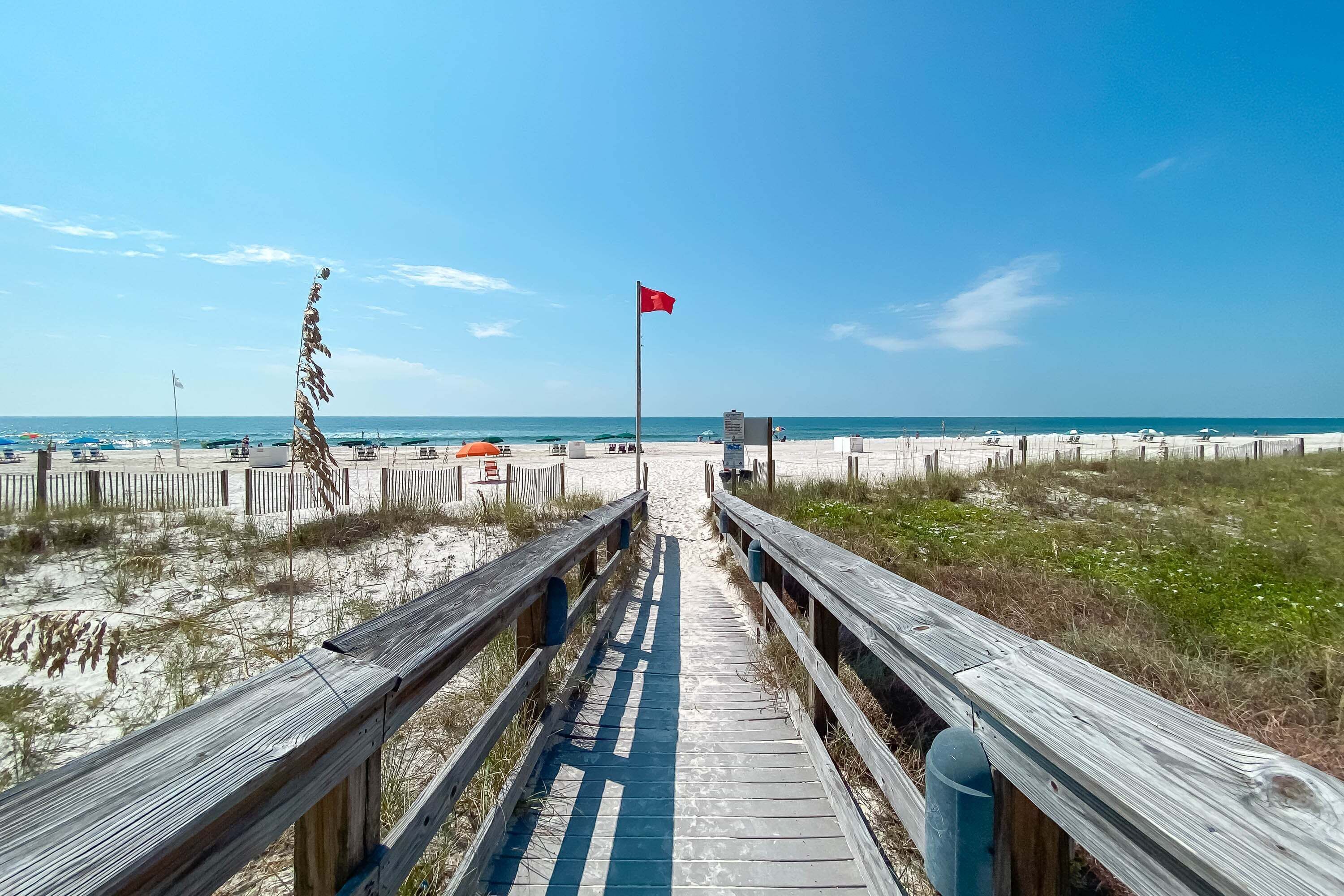 Ein Holzsteg führt zu einem Strand mit einer roten Flagge, Sanddünen und einem klaren, blauen Himmel.