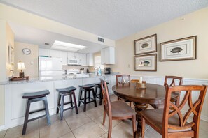 Cocina con armarios blancos, microondas y cafetera. Hay cuatro taburetes de madera y una mesa de comedor de madera con sillas.