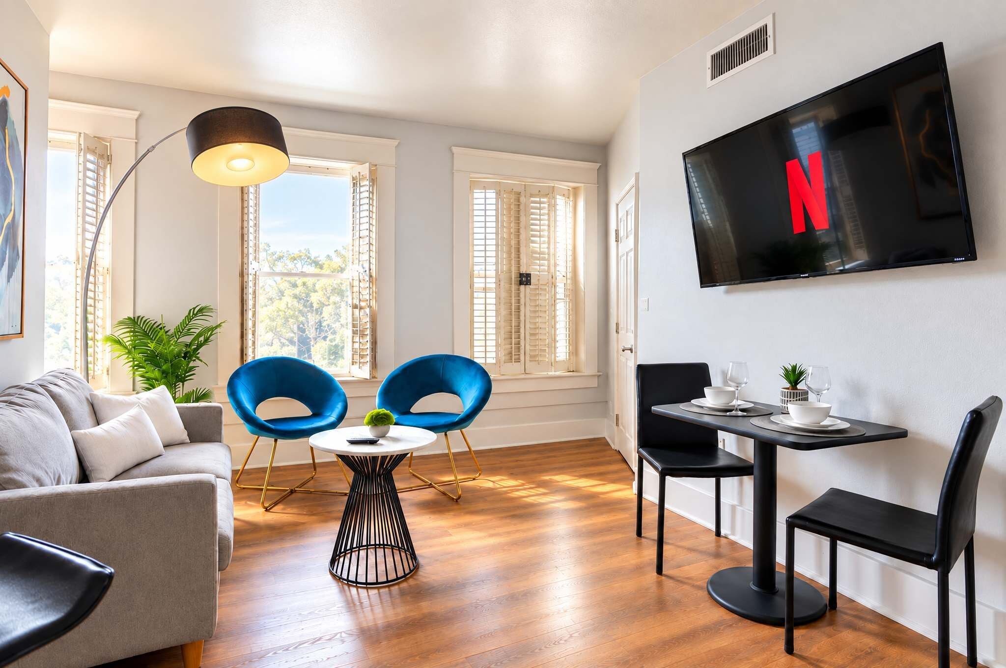 Ein modernes Wohnzimmer mit einer Couch, zwei blauen Sesseln, einem kleinen runden Tisch, einem an der Wand befestigten Fernseher und einem Esstisch mit Stühlen.