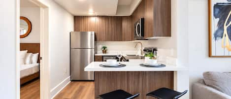 A modern kitchen with a built-in refrigerator, microwave, and wooden cabinets. There are two black bar stools at the island, and a dining table with plates and glasses.