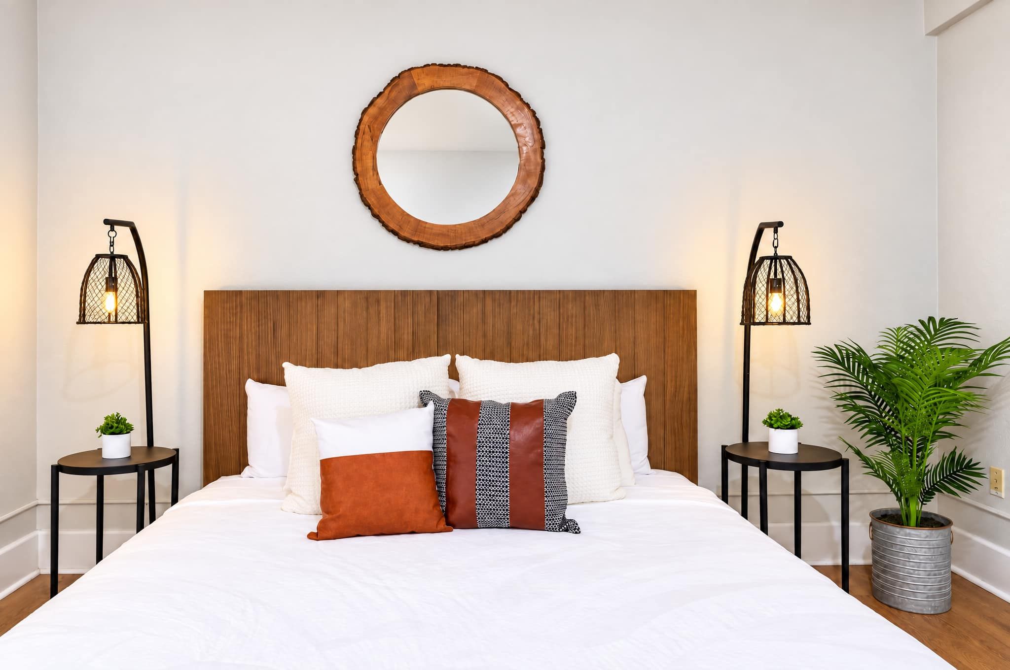 A neatly made bed with a wooden headboard, two bedside tables with lamps, a round mirror, and a potted plant.
