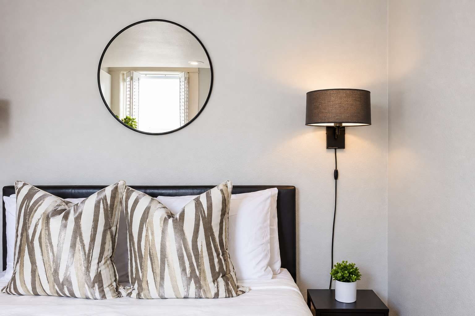 Quarto de hotel com uma cama, um espelho redondo, uma luminária na parede e um vaso pequeno com planta em uma mesa de cabeceira.