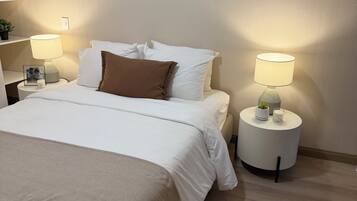 Una cama bien hecha con ropa blanca y un plaid marrón, dos lámparas de noche, y una mesita de noche con una planta y un marco de fotos.
