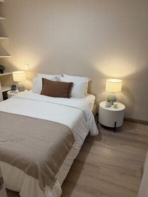 A neatly made bed with white bedding and a brown throw, two bedside lamps, and a small bedside table with a plant and a picture frame. - Vrbo Property (Jesús María)