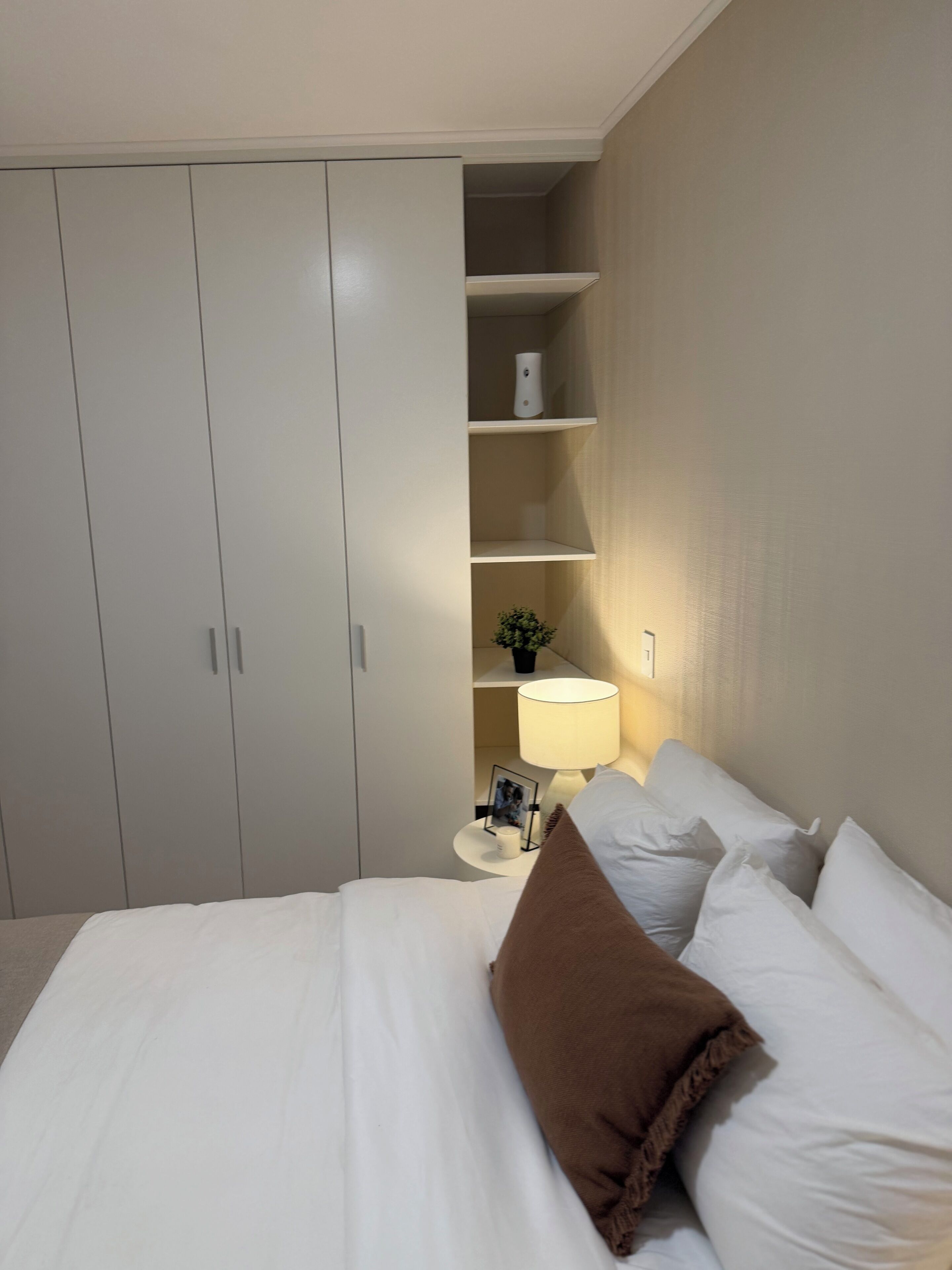 Un dormitorio con una cama, almohadas, una mesita de noche, una lámpara, un estante con una planta y un armario.