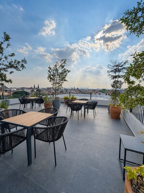 Een terras buiten met tafels en stoelen, planten in potten en uitzicht op een stadsbeeld.