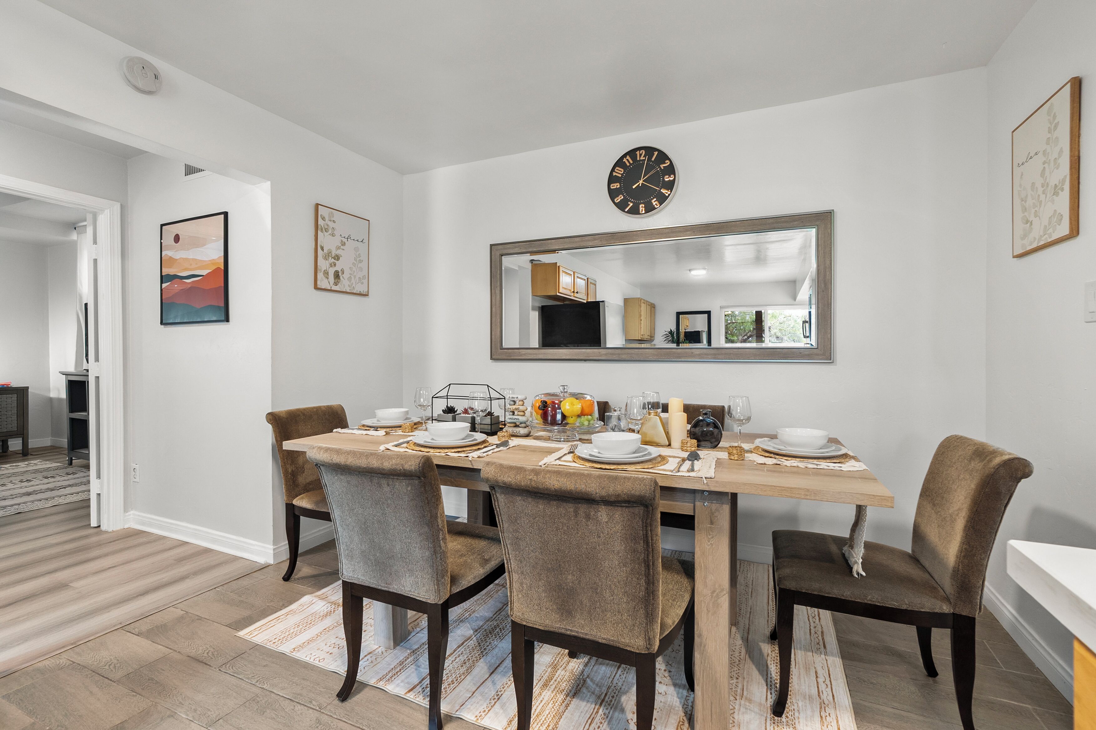 A dining area with a wooden table set for four, surrounded by upholstered chairs. A large mirror hangs above the table, reflecting the room.