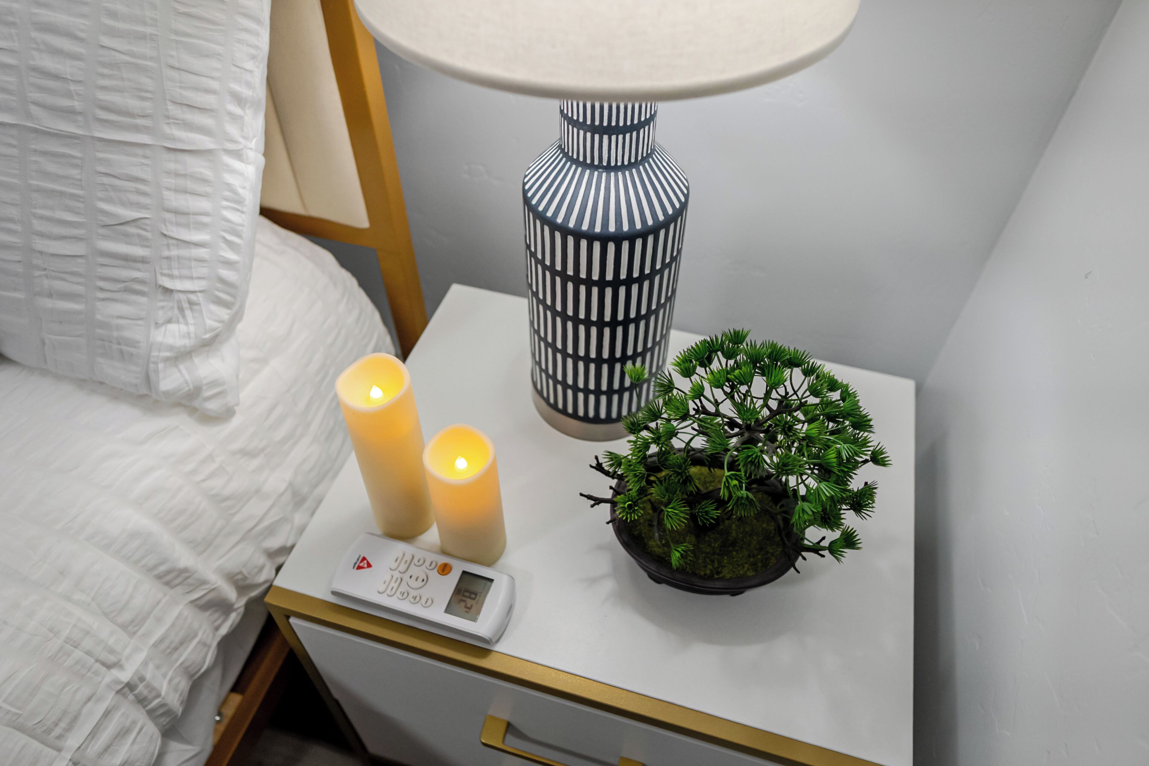 A neatly made bed with white bedding, a bedside table with a digital thermometer, two lit candles, a blue and white patterned lamp, and a small potted plant.