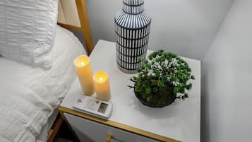 Una cama bien hecha con ropa de cama blanca, una mesita de noche con termómetro digital, dos velas encendidas, una lámpara con estampado azul y blanco, y una pequeña planta en maceta.