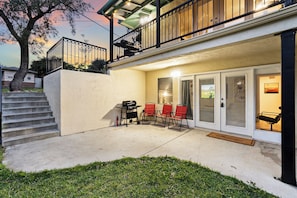 A covered patio with a grill and red chairs, a staircase leading to a balcony, and a door with a glass panel.
