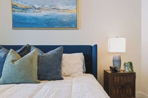 Een slaapkamer met een bed, kussens, een nachtkastje, een lamp en een schilderij aan de muur.