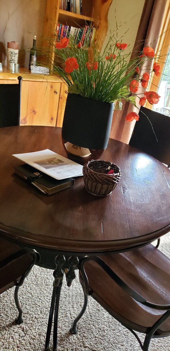 Een ronde houten tafel met een zwart metalen onderstel, een potplant met rode bloemen, een klein geweven mandje en een boek.