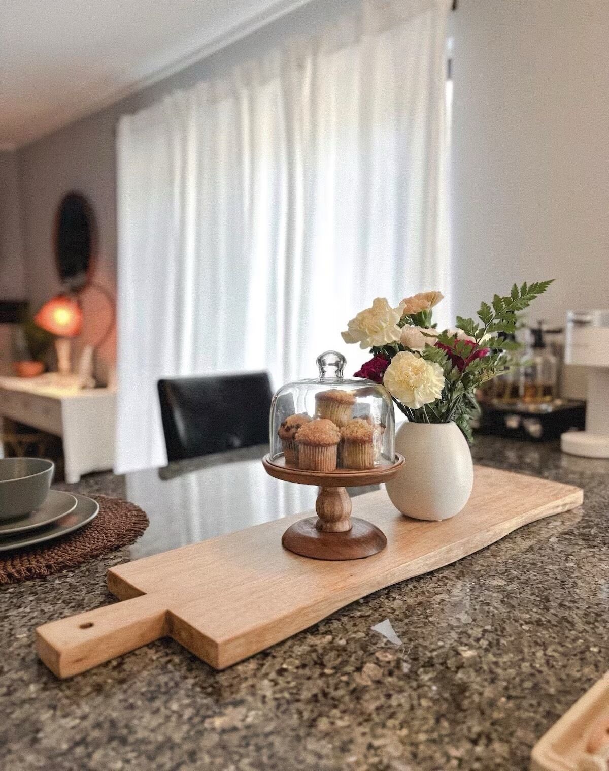 A kitchen counter with a glass display case containing muffins, a vase with flowers, and a wooden cutting board.