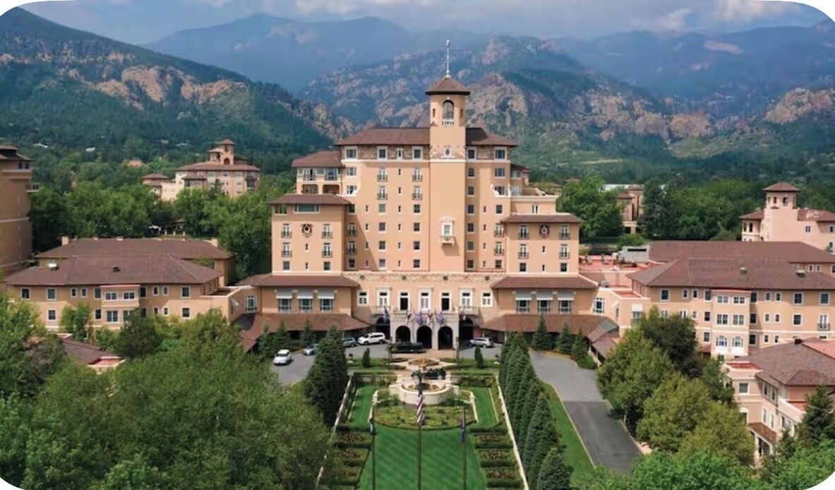 A large, multi-story building with a central tower, surrounded by lush greenery and mountains in the background.