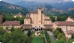 Un grand bâtiment à étages, doté d’une tour centrale, entouré d’une végétation luxuriante et avec des montagnes en arrière-plan.