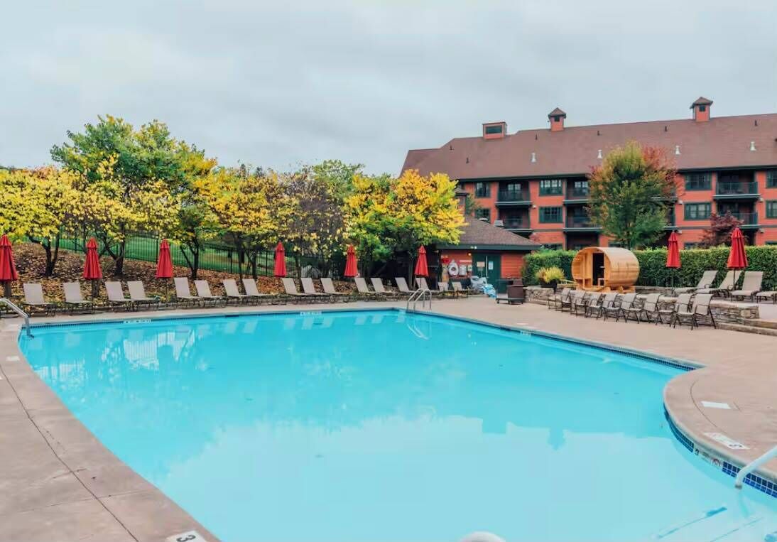 Une piscine entourée d’arbres et d’un bâtiment doté de nombreuses fenêtres, avec des chaises longues et des parasols à proximité.