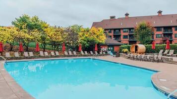Une piscine entourée d’arbres et d’un bâtiment doté de nombreuses fenêtres, avec des chaises longues et des parasols à proximité.