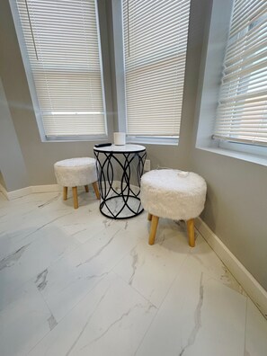 A room with a marble floor, two white furry ottomans, a black metal table, and a window with blinds.