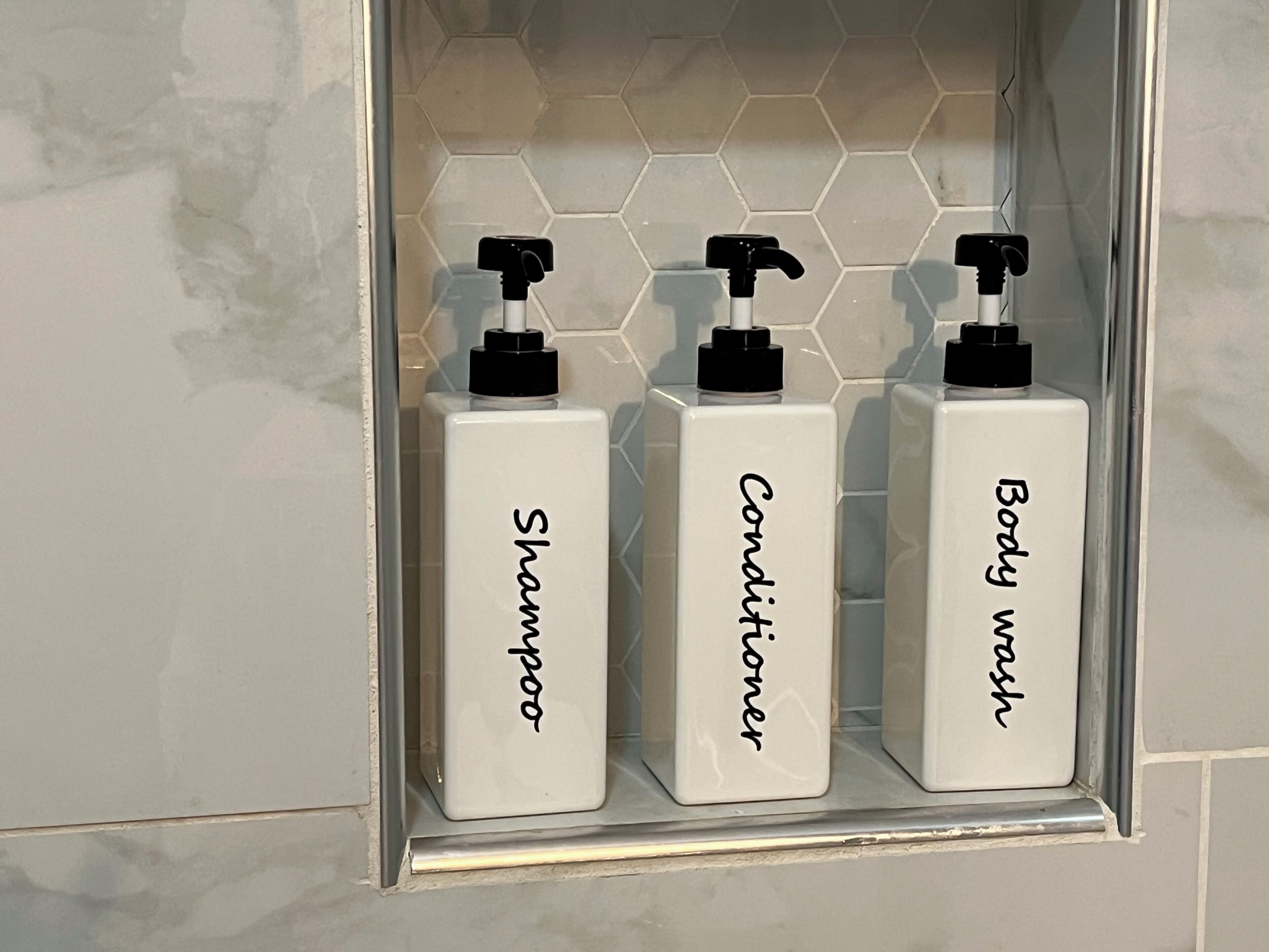 Trois flacons blancs avec des pompes noires, étiquetés « Shampoing », « Après-shampoing » et « Gel douche », sont disposés dans une salle de bain.