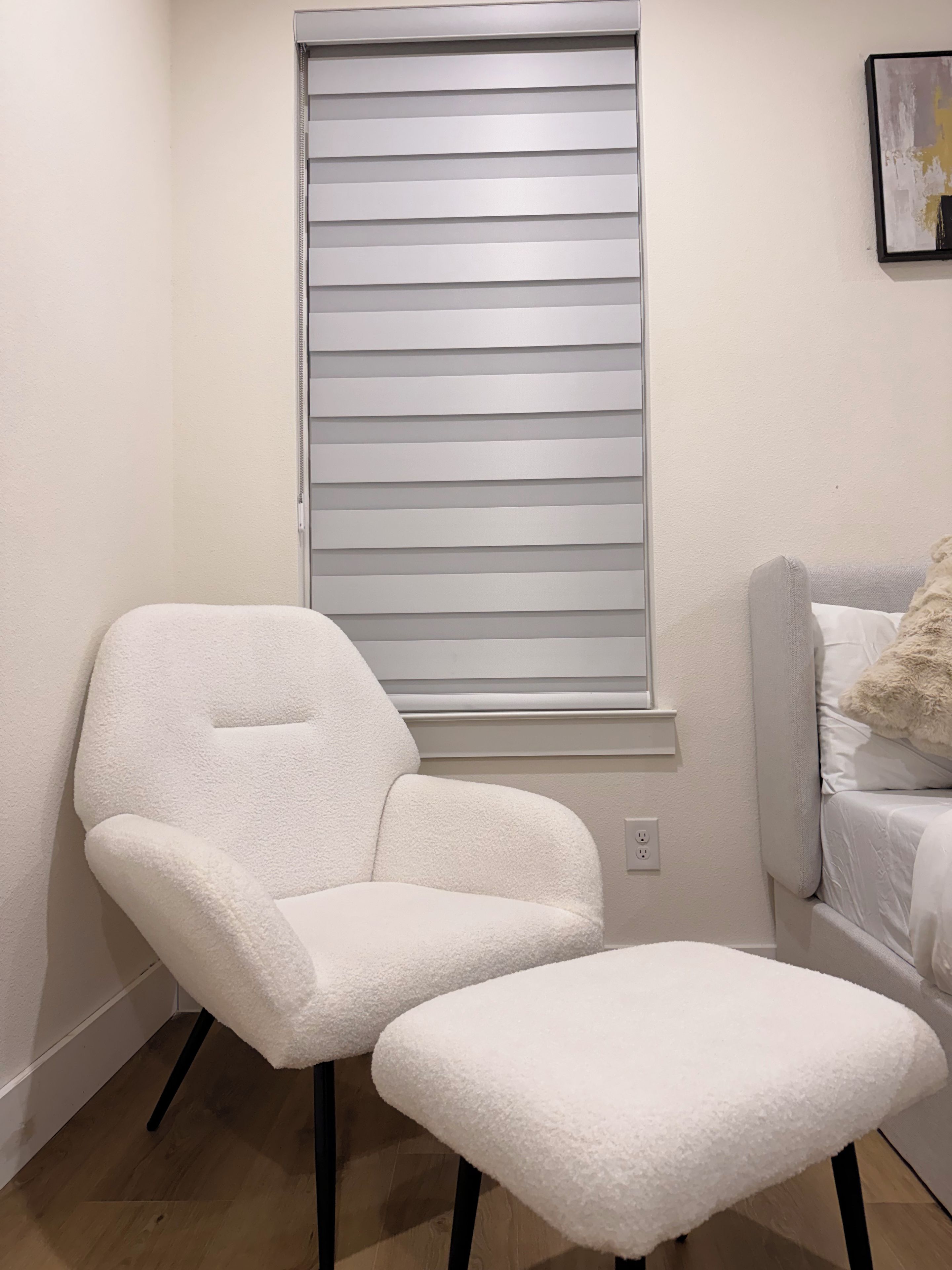 A room with a white chair and ottoman, a window with horizontal blinds, and a bed with white bedding.